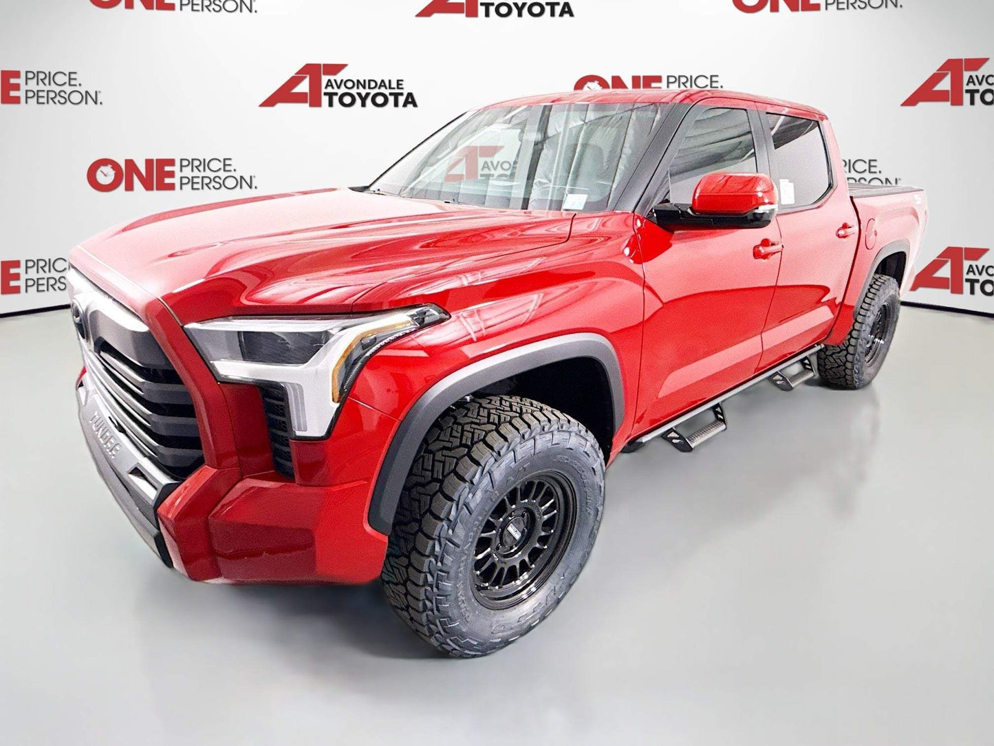 Red 2025 Toyota Tundra pickup truck with lifted suspension and off-road tires in showroom.